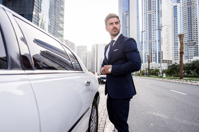 A businessman in a black suit alongside a white limousine on an empty street with tall buildings on the background.
